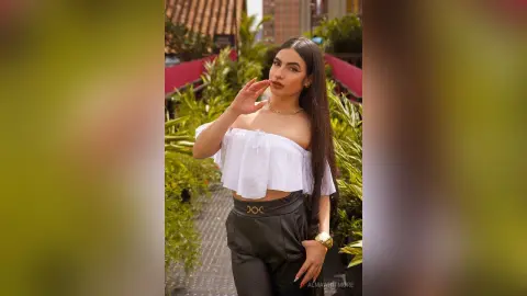 AlmaWhitmore poses outdoors wearing a white off-the-shoulder crop top and black pants, with her long dark hair down.