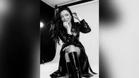 Glamorous woman in a black leather trench coat and tall boots posing in a studio setting.