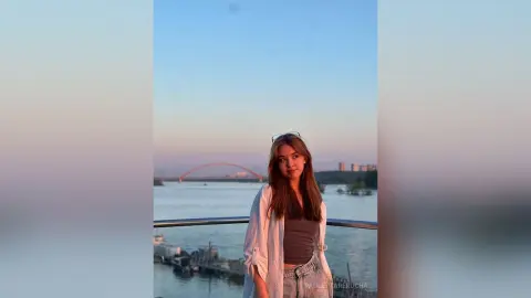 PaulettaRerucha stands on a balcony overlooking a river and a red bridge, wearing a grey tank top and a white open shirt.