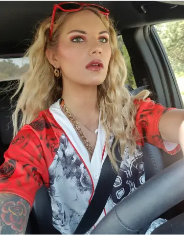 GoddessBiancaBelle with long blonde hair and red eyeshadow wears a red and white patterned shirt while sitting in a car.