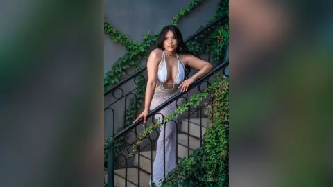 DayanaHille poses on a staircase wearing a white plunging halter neck top and a long white skirt, leaning against a black metal railing surrounded by green ivy.