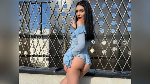 Young woman in a light blue ruffled mini dress posing outdoors against a metal lattice fence in bright sunlight.
