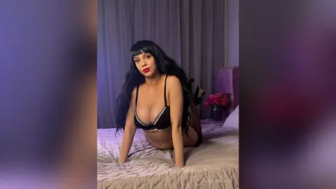 Young woman with long black hair in a black bikini posing on a bed in a moody bedroom setting.