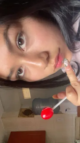 Close-up portrait of a young woman with long dark hair holding a red lollipop in a bright bathroom setting.