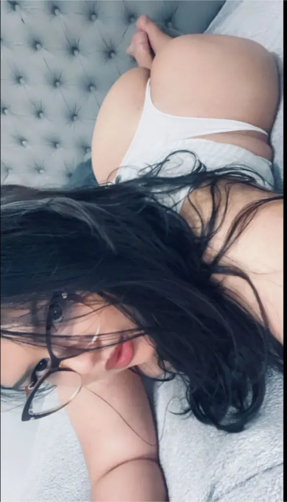 Young woman with long dark hair and glasses wearing a white button-down shirt in a soft indoor setting.
