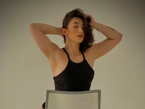 Katt_Henao poses with her hands behind her head, wearing a black halter top against a plain white wall.