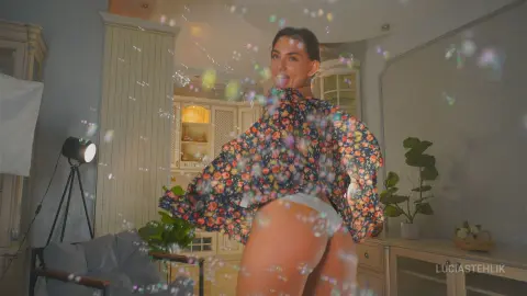 Cheerful woman in a floral print blouse dancing amidst floating bubbles in a bright, cozy living room.