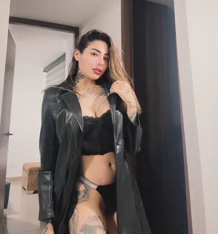 Tattooed woman in a black leather coat and lingerie posing confidently in a modern minimalist room.