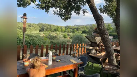 Blonde woman sitting at a wooden table on a rustic outdoor terrace surrounded by lush green trees.