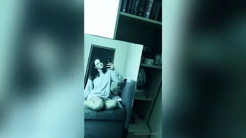 Young woman in a cozy white sweater taking a moody mirror selfie in a dimly lit bedroom.