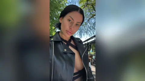 Young woman in a black leather jacket and strappy top posing outdoors in a lush green park setting.