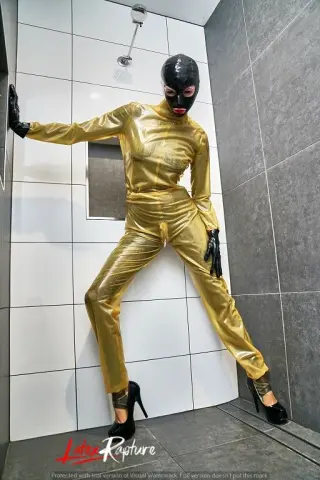 LatexRapture wears a shiny gold bodysuit and black gloves, posing in a white tiled shower stall while wearing black high heels.