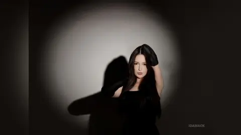 Elegant woman in a black dress posing under a dramatic spotlight in a moody, dark studio setting.