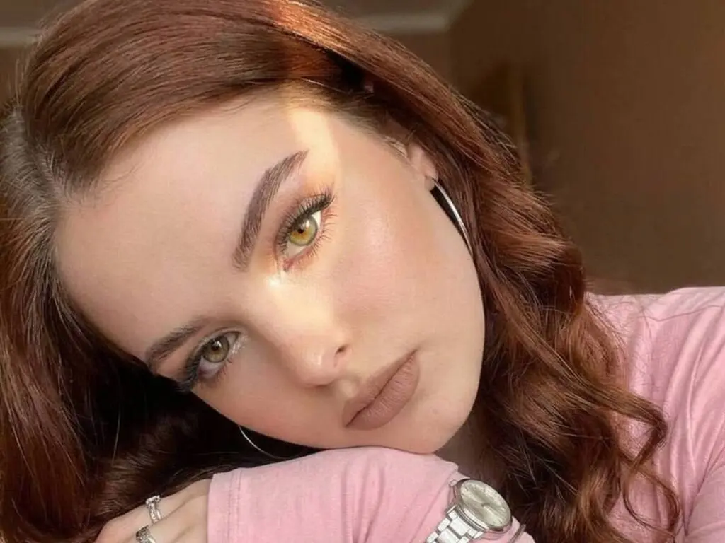 Close-up portrait of a woman with wavy red hair and glowing makeup wearing a pink top in soft sunlight.