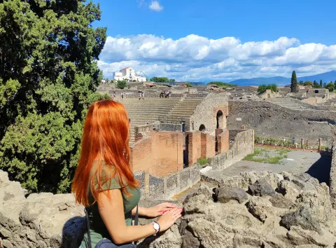 LeilaBlissXO with long red hair, wearing a green t-shirt, looks out over the stone ruins of an ancient city under a blue sky.