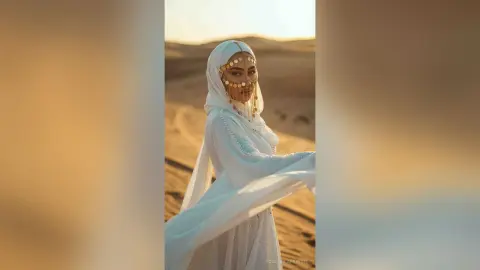 Elegant woman in flowing white ethereal gown and gold headpiece posing in a dreamy desert landscape at sunset.