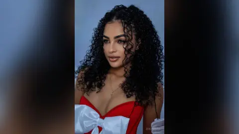 Young woman with curly dark hair wearing a red strapless top with a white bow in a soft studio setting.