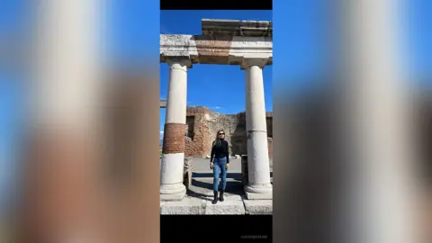 Woman in black long-sleeve shirt and jeans standing by ancient stone ruins under a bright blue sky.