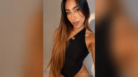 VictoriaFontaine poses in a black one-shoulder bodysuit, showing her long brown hair and cleavage against a plain background.