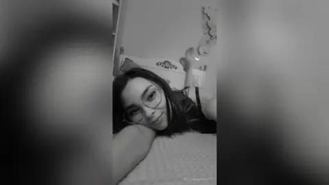 Young woman with glasses relaxing on a bed in a cozy, moody black and white indoor setting.