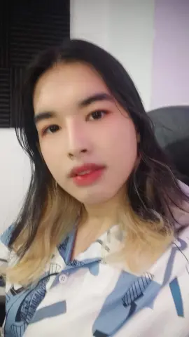 Young woman with long dark hair wearing a white and blue patterned shirt in a cozy indoor setting.