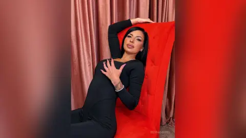 ReynaMorel poses in a tight black long-sleeve bodysuit while leaning back against a bright red velvet chair.