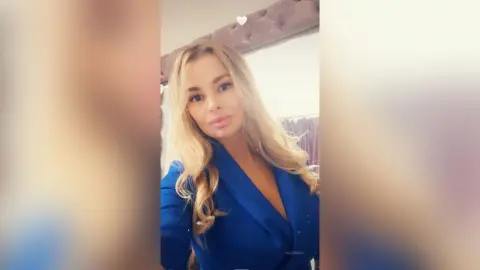 MilenaBia poses with long blonde hair wearing a deep blue v-neck top or blazer.