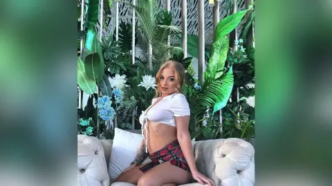 JeniferMoon — JeniferMoon sits on a white sofa wearing a white crop top and a red plaid mini skirt amidst green tropical plants.