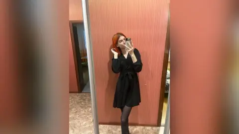 Red-haired woman in a black long-sleeve dress taking a mirror selfie in a modern room.