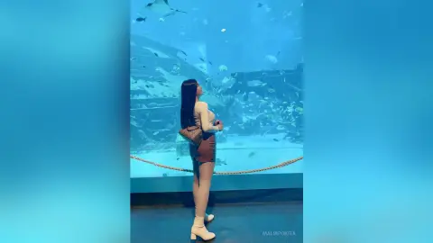 Woman in a beige top and brown skirt gazing at fish inside a blue aquarium tunnel.