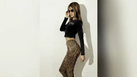 Stylish woman in a black long-sleeve crop top and leopard print leggings posing against a white wall.