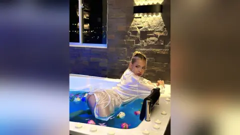 Young woman in a white satin robe relaxing in a luxury bathtub filled with rose petals at night.
