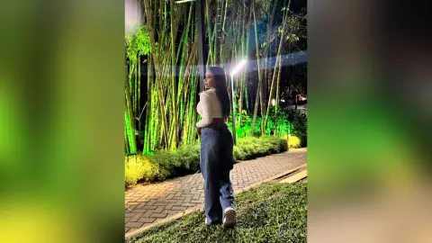MiaKorz stands outdoors at night in a white long-sleeve top and blue jeans, posing next to a thick bamboo forest and a brick walkway.