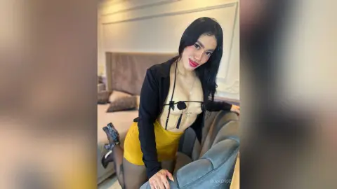 Smiling woman in a black cardigan and yellow mini skirt posing in a modern bedroom setting.