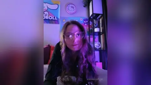 Young woman with long wavy hair and glasses in a purple lit bedroom with aesthetic posters.