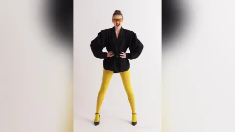 Cheerful woman in black blazer and bright yellow leggings posing against a white studio background.