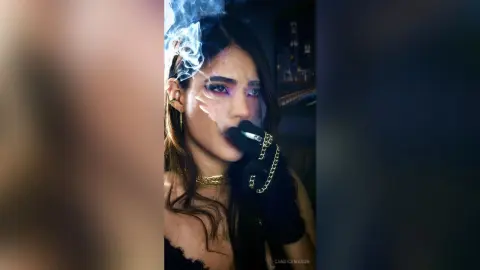 Moody portrait of a woman with smoky eye makeup and gold jewelry exhaling smoke in a dark setting.