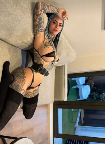 Tattooed woman in black lingerie and stockings relaxing on a grey sofa in a bright modern room.