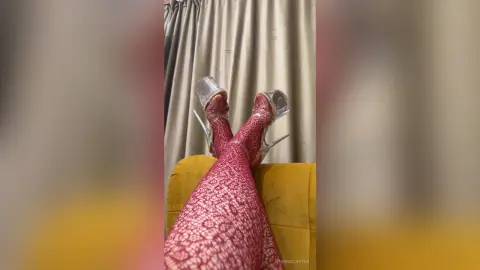 Close-up of legs in red lace patterned tights and clear high heel sandals resting on a yellow velvet sofa.