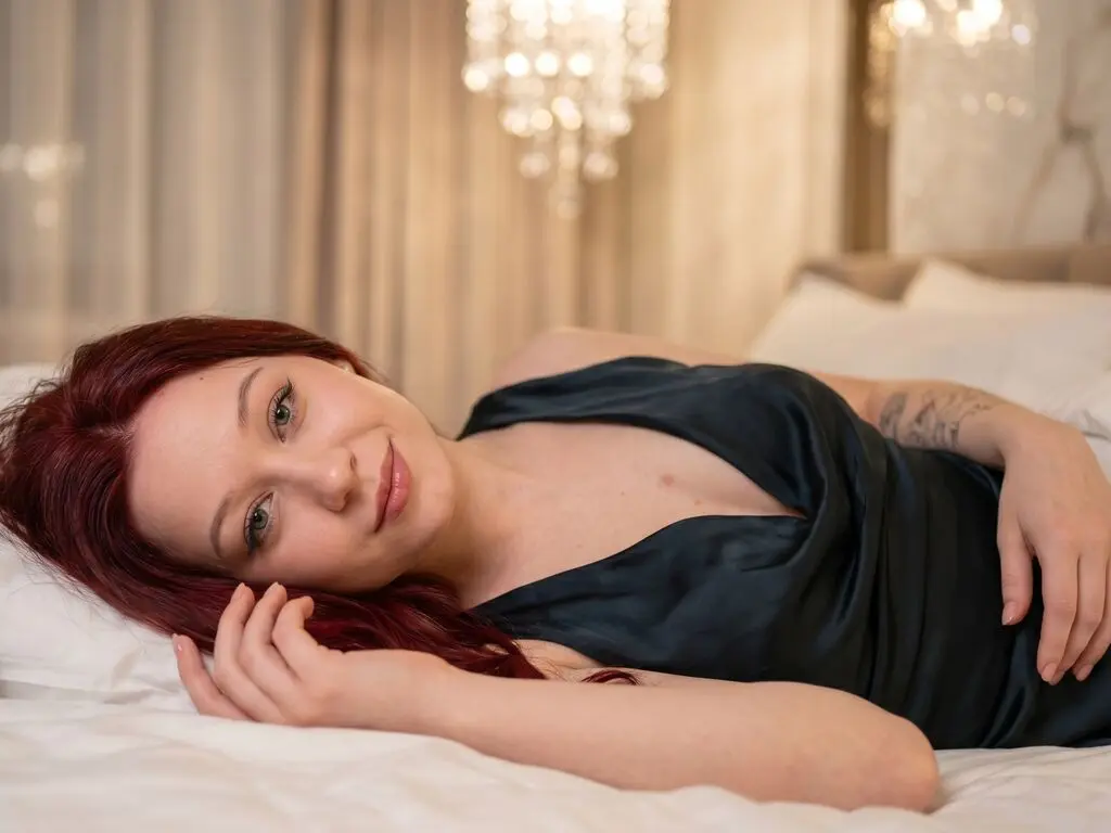 Redheaded woman in a dark silk dress relaxing on a white bed in a warm, cozy bedroom setting.