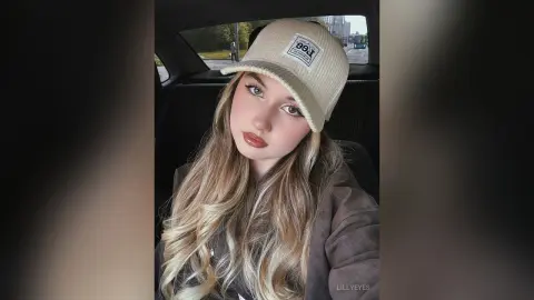 LillyEyes wearing a tan baseball cap and a brown jacket, posing with long blonde hair in a car seat.