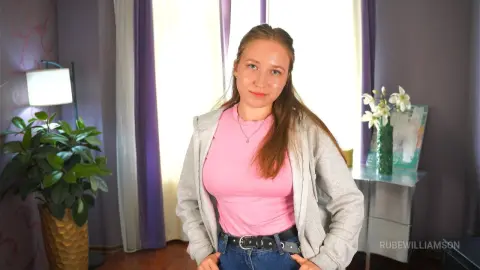 RubeWilliamson wears a pink t-shirt and grey zip-up hoodie with blue jeans, standing in a room with a plant and purple curtains.