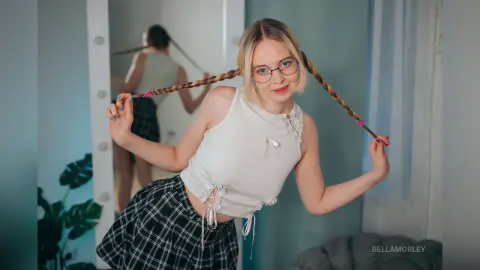 Cheerful blonde woman in a white sleeveless top and plaid skirt posing with long braided hair in a bright room.