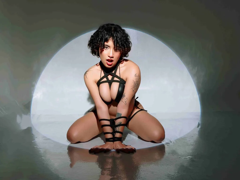 DixieLyons — DixieLyons poses in a black corset and halter top while leaning against a metal pole in a dark club setting.