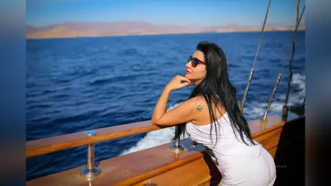 MissyJolie leans against a wooden boat railing overlooking the blue ocean, wearing a white tank top and red sunglasses.