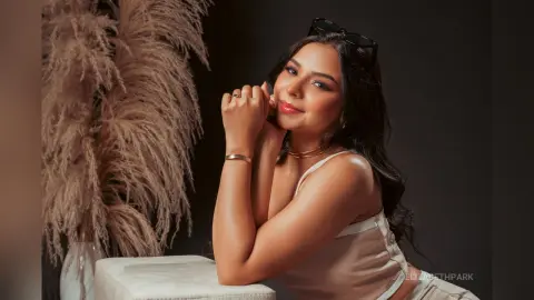 Smiling young woman in a white satin top posing elegantly next to decorative pampas grass in a warm studio setting.