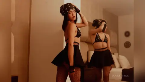 Young woman in black bikini top, skirt, and headscarf posing in a warm-toned bedroom setting.