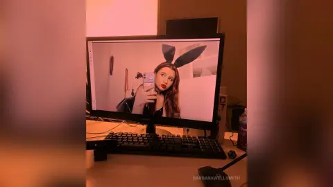 Young woman with bunny ears taking a selfie on a computer monitor in a warm, moody room setting.