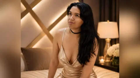 Smiling woman in a gold satin slip dress sitting on a bed in a warm, dimly lit bedroom.