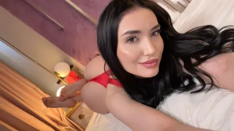 Smiling woman with long black hair relaxing in red lingerie on a white bed in a cozy bedroom setting.